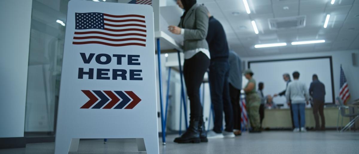 This image shows a "Vote Here" sign in the foreground with the American flag at the top and arrows pointing forward. In the background, a diverse group of people is standing in line at voting booths, filling out ballots inside a well-lit polling station. 